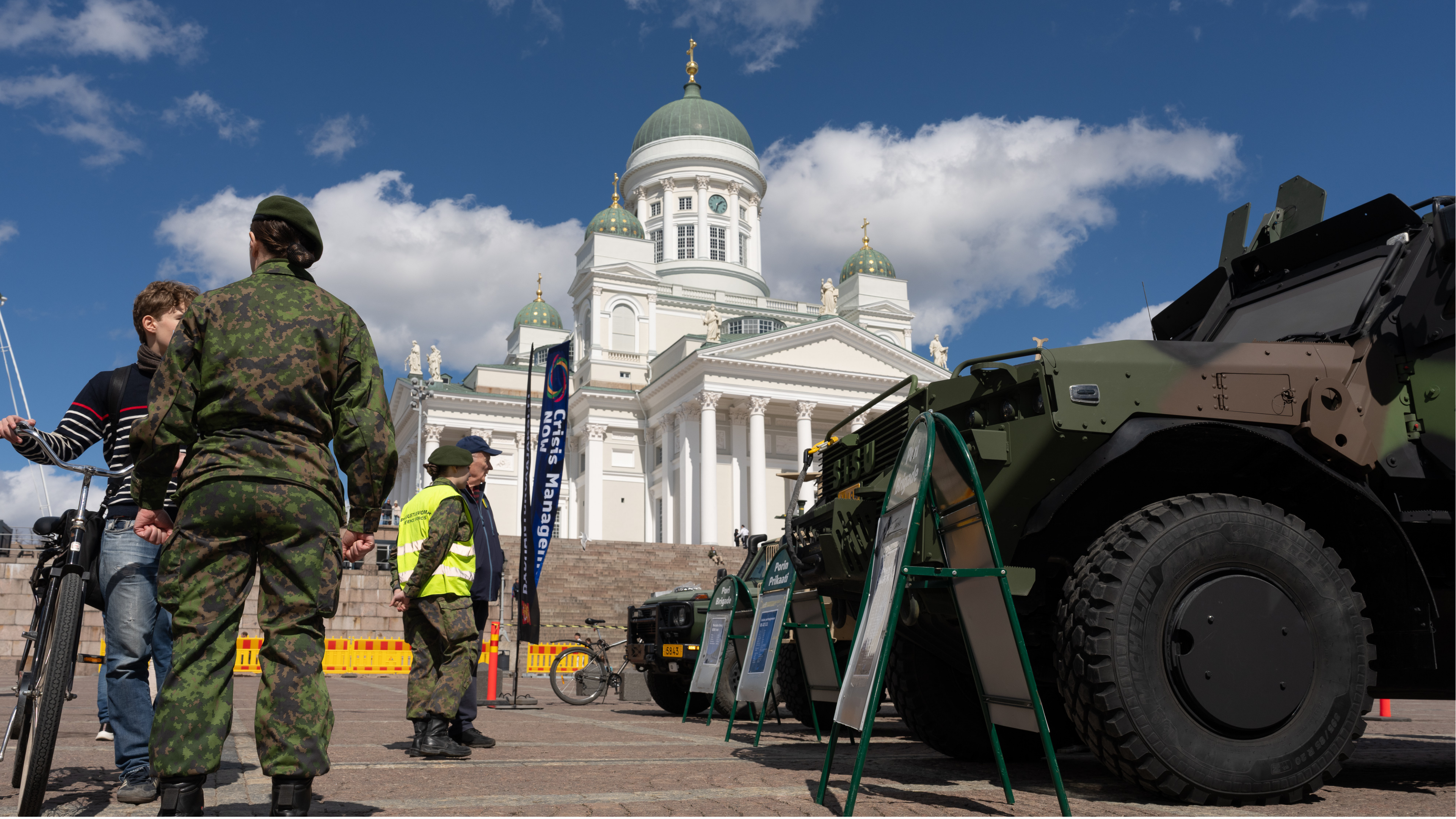 Senaatintorilla seisovia sotilaspukuisia henkilöitä keskustelemassa ulkona avoimen yleisötapahtuman yhteydessä; oikealla etualalla suuri vihreä sotilasajoneuvo infotauluineen ja tikkaineen, taustalla Helsingin tuomiokirkko valkoisine pylväineen ja vihreine kupoleineen sinistä taivasta vasten.