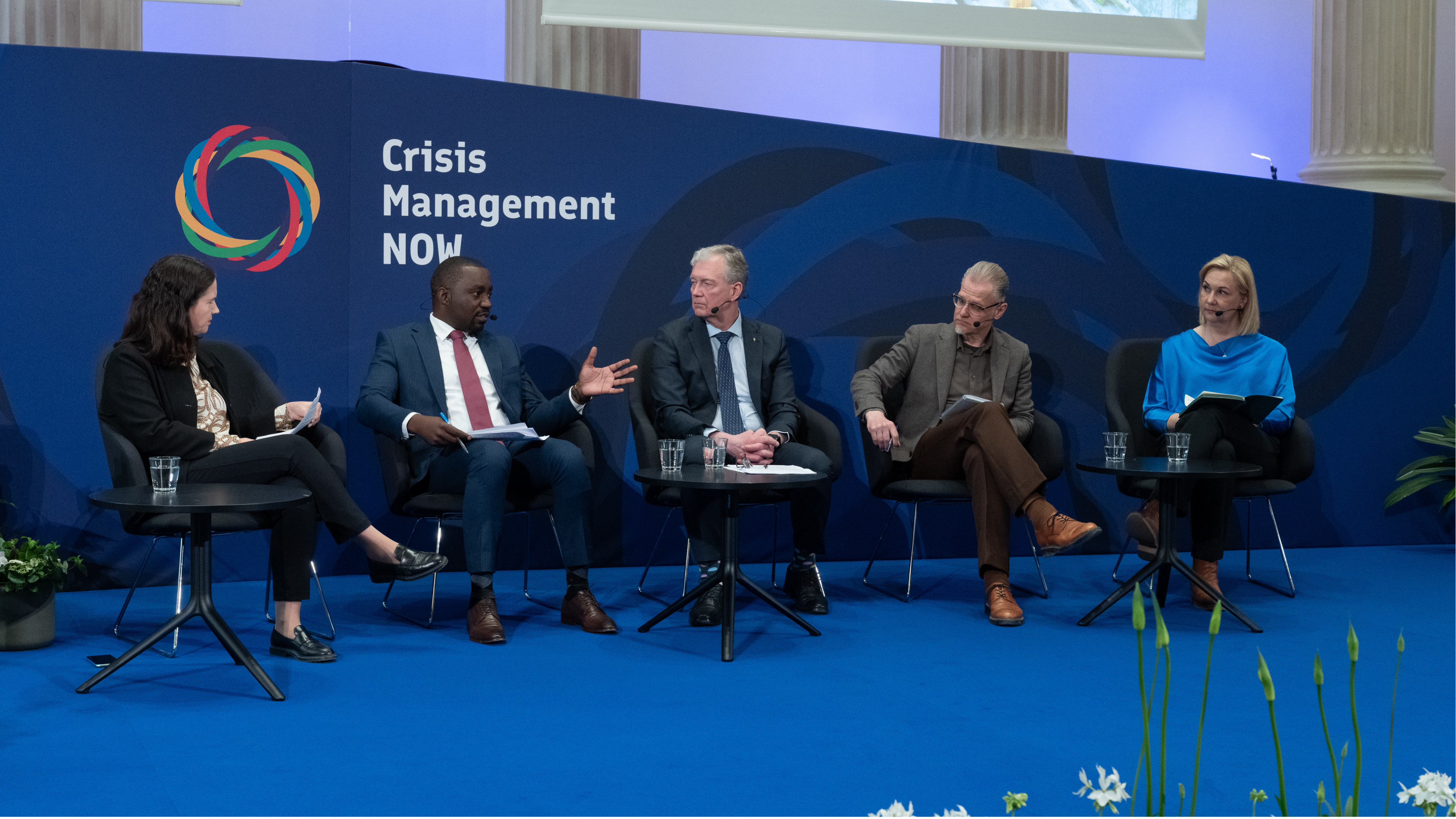 Usean hengen paneelikeskustelu lavalla: keskustelijat istuvat tuoleilla pienien pöytien ääressä, joilla on vesilaseja ja muistiinpanoja; taustalla suuri sininen seinä, jossa näkyy teksti ”Crisis Management NOW” ja värikäs ympyrälogo, etualalla sininen matto ja istutuksia.