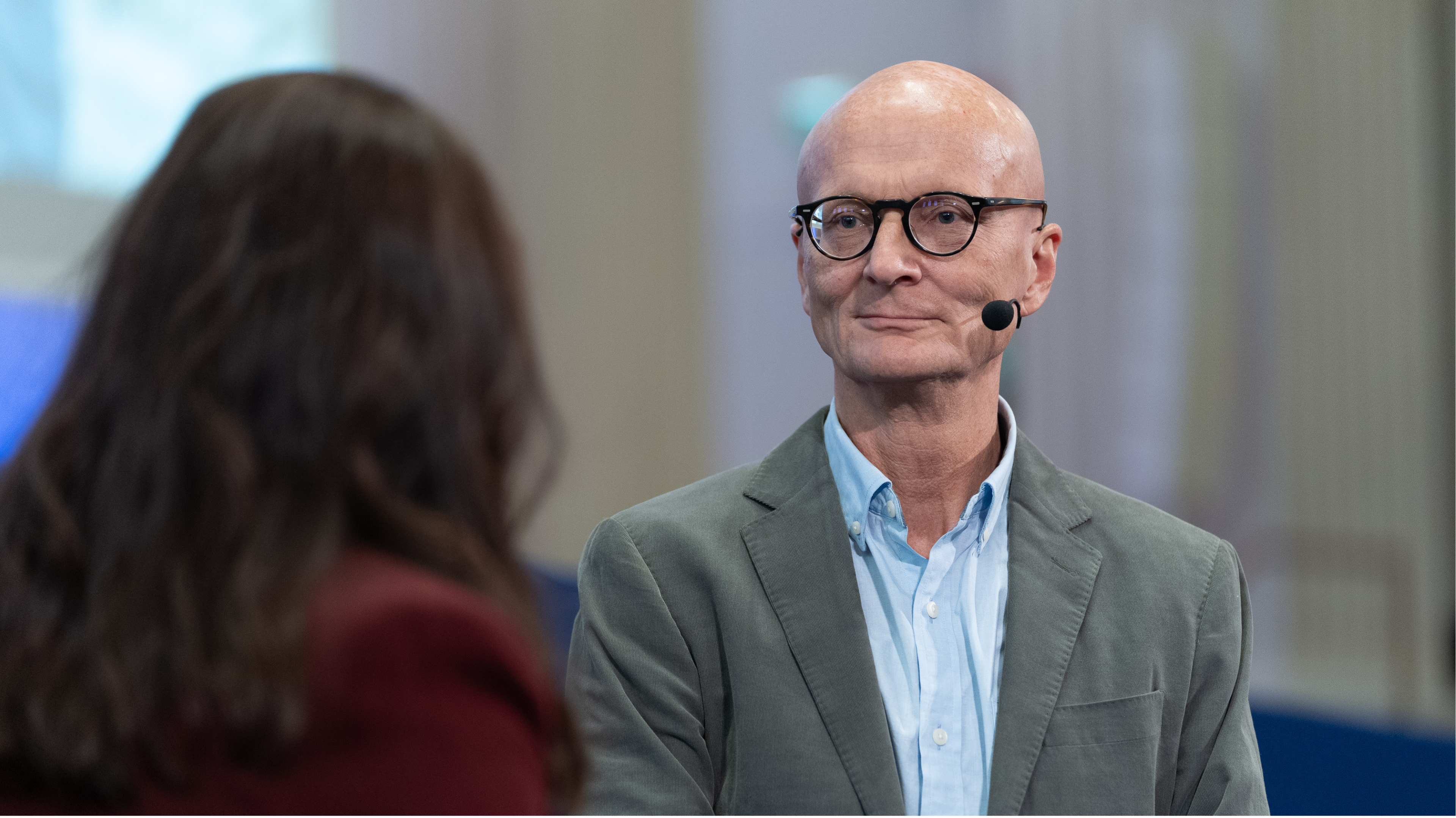 Lähikuva puhujasta sisätilassa: henkilö käyttää korvamikrofonia ja katsoo keskustelukumppania kohti; keskustelukumppani näkyy epätarkkana etualalla, ja tausta on hillityn valaistu ja neutraalin värinen.