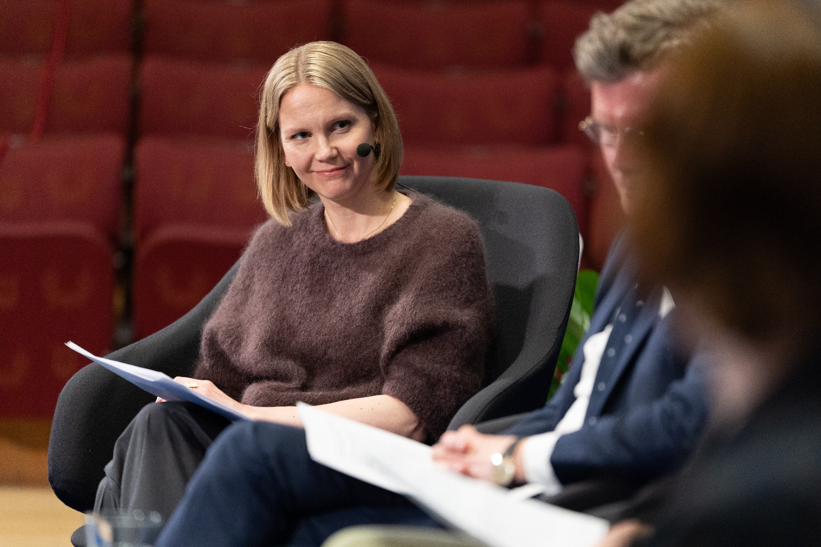 Nainen osana paneelina hymyilee ja katsoo muihin panelisteihin joita kuvassa näkyy kaksi kappaletta. 