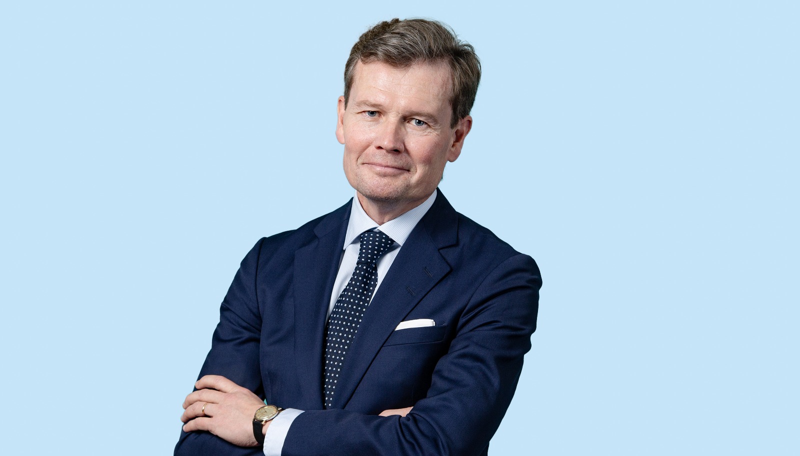 Mies puku päällä katsoo kameraan, kädet rinnan päällä ristissä