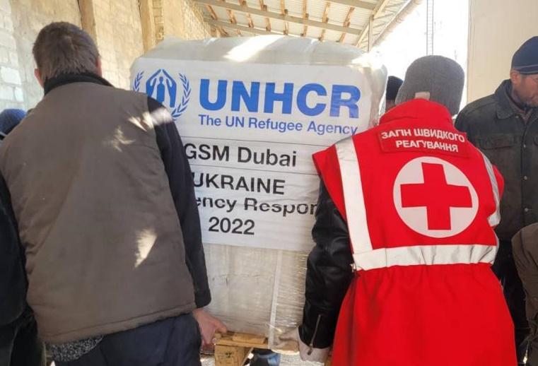 Two people carrying a big package of UNCHR supplies to Ukraine. One of them is wearing a vest with a Red Cross emblem.