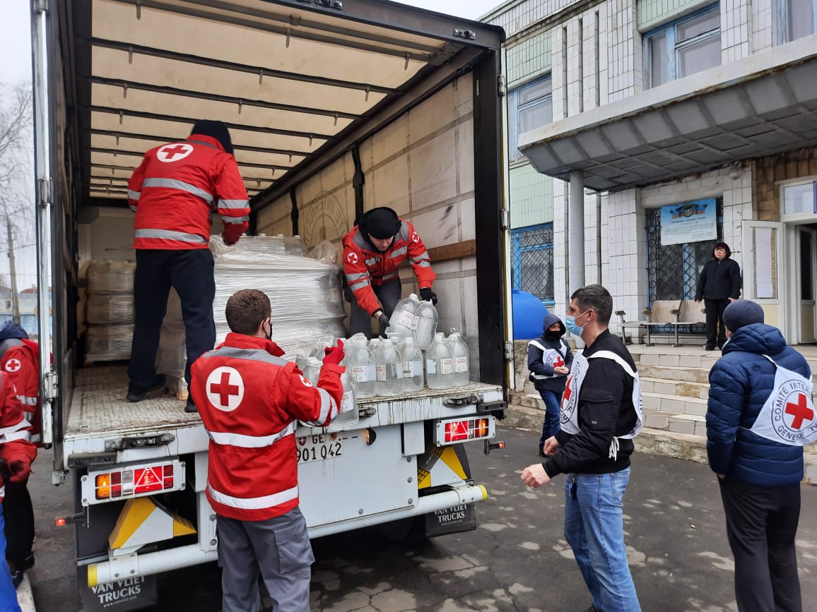 People wearing jackets with a symbol of the Red Cross are handing out water cans from a truck.