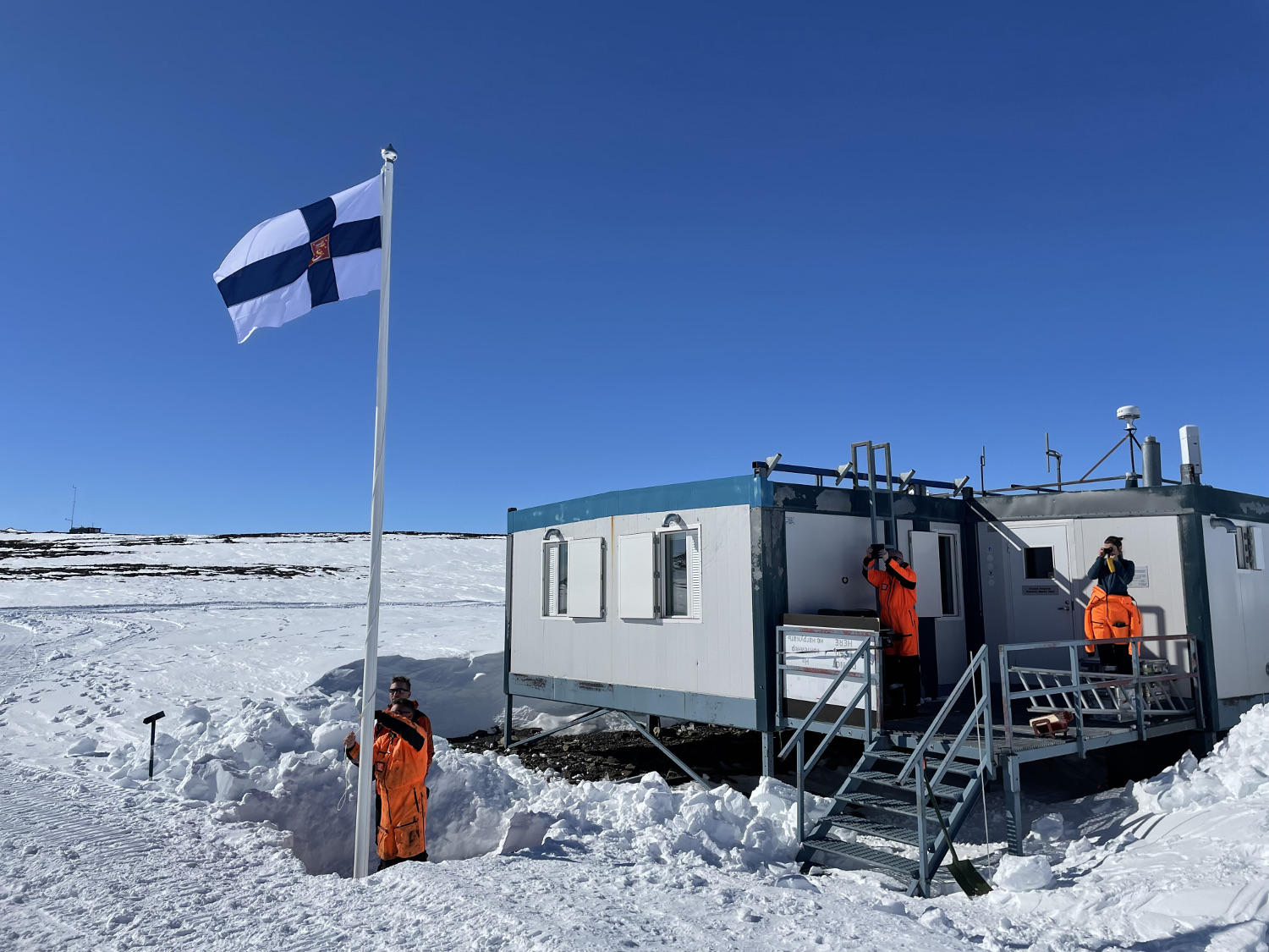 Mies seisoo lumihangessa ja nostaa Suomen lippua salkoon. Taustalla on Suomen tutkimusaseman parakkimainen rakennus. Taivas on kirkkaan sininen, ympäristö on tasaista, horisontti näkyy kaukana.
