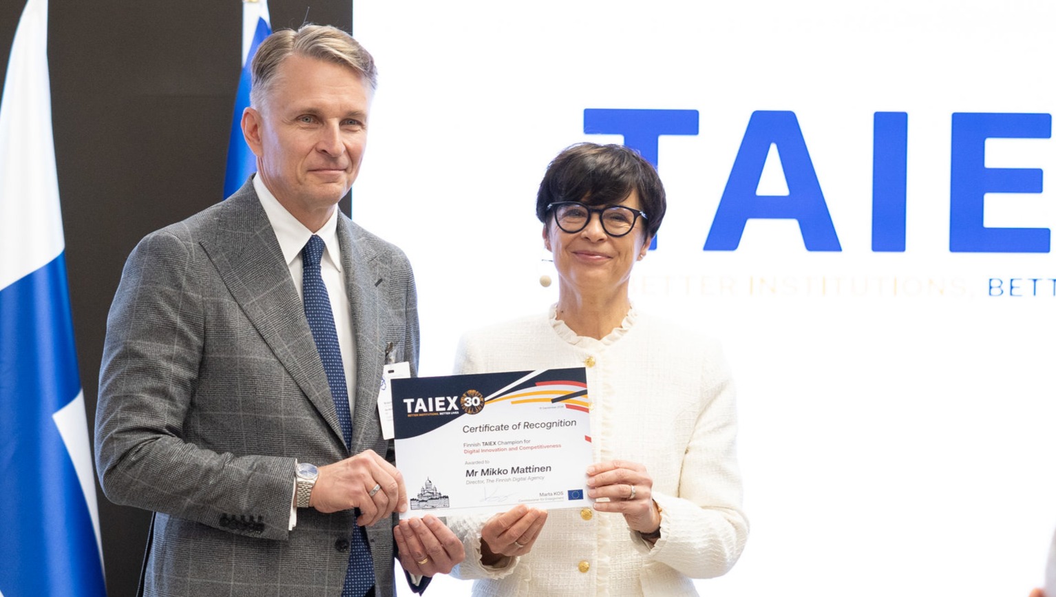 Mikko Mattinen vastaanottaa tunnustuskirjan Marta Kosilta TAIEX-ohjelman 30-vuotisjuhlassa. Taustalla näkyvät Suomen liput ja TAIEX 30 -juhlagrafiikkaa.