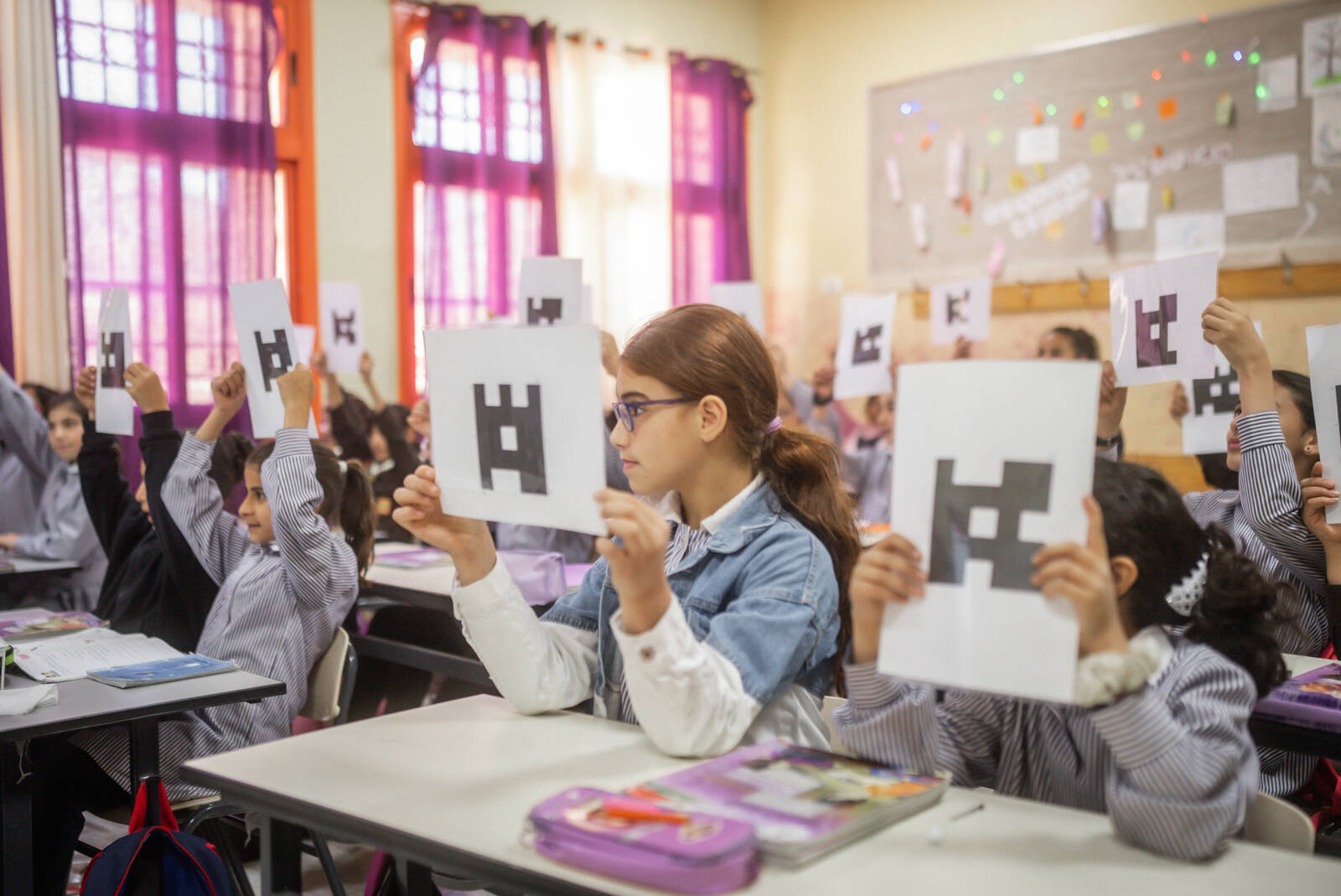 Koululaiset istuvat pulpeteissa ja näyttävät papereita opettajalle. Kuva: Mosadaq Muhannad Qadan / Studio Havana