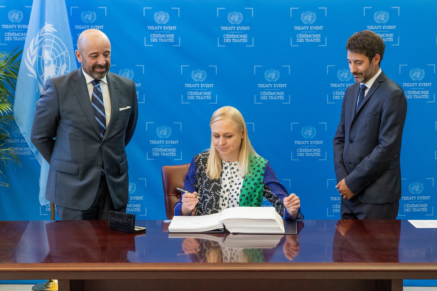 Ministeri Elina Valtonen istuu pöydän ääressä kynä kädessään ja edessään avattu kirja. Kaksi mieshenkilöä seisoo pöydän molemmin puolin. Taustalla YK-lippu.