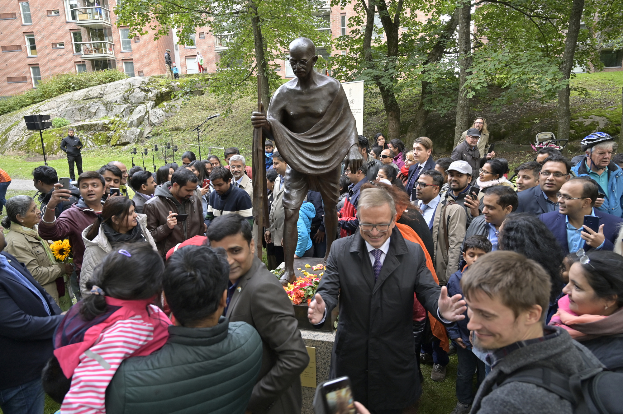 Patsas esittää Mahatma Gandin luonnollisen kokoisena pronssifiguurina. Patsaan ympärillä tilaisuuteen osallistuneita ihmisiä.