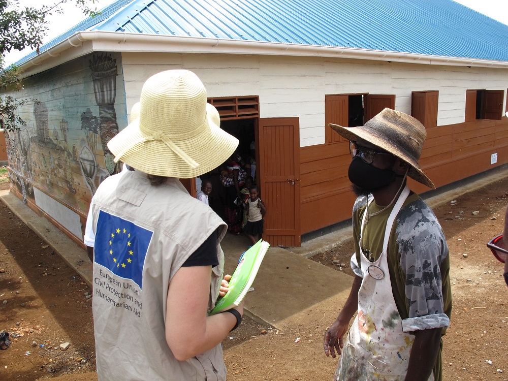 An aid worker pictured has a big cylinder hat and an EU flag on her back that reads Humanitarian Aid. Opposite her stands a black man with a cylindrical hat and a mask. Behind them, children peek out the door.