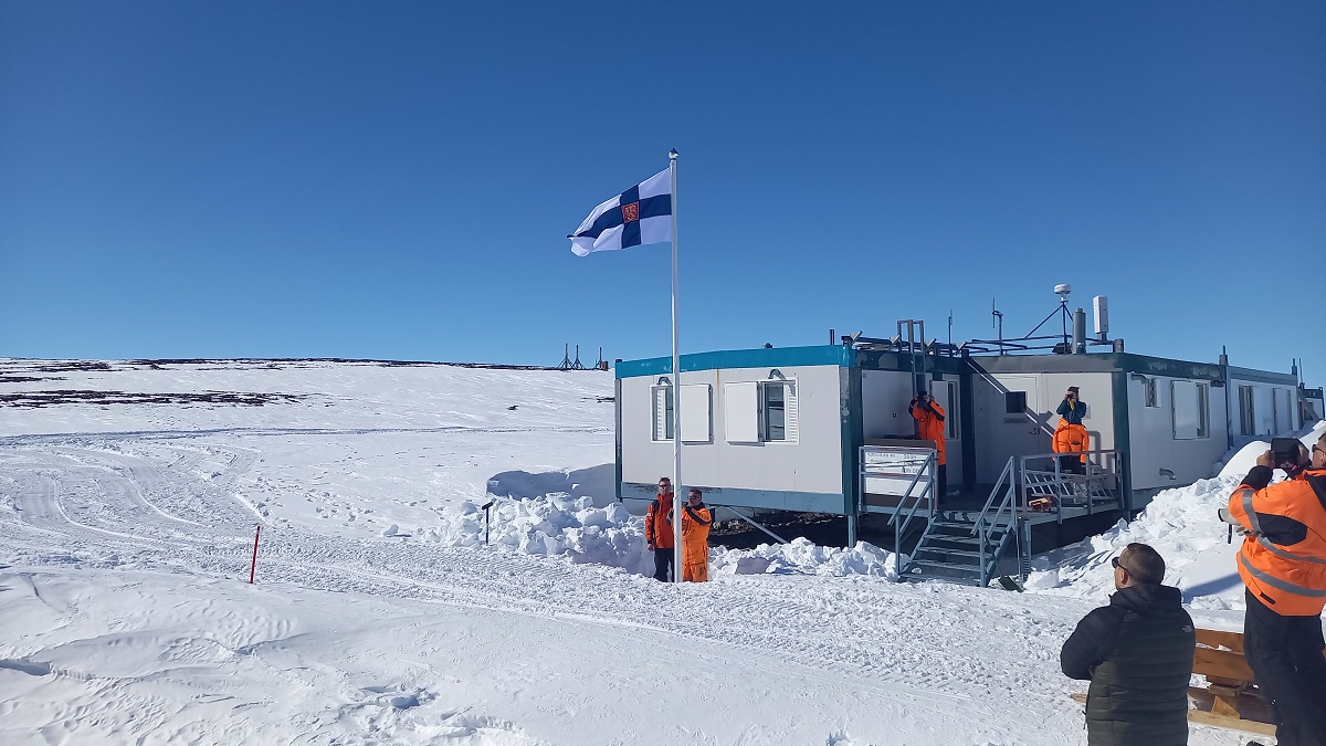 Kuvassa luminen maisema Etelämantereella, pari parakkirakennusta ja keskellä lipputanko, missä Suomen lippu.