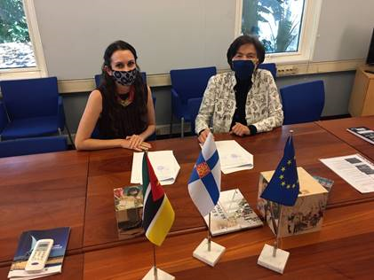 Pöydän ääressä istuu kaksi naista maskit kasvoilla. Mosambikin, Suomen ja EU:n liput edustalla.